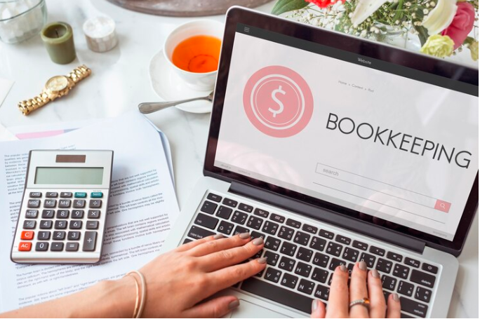 A person at a messy desk typing on a laptop with the word bookkeeping on the laptop screen.