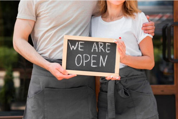 A couple standing close to each other and they are both holding a chalkboard sign that says "we are open".